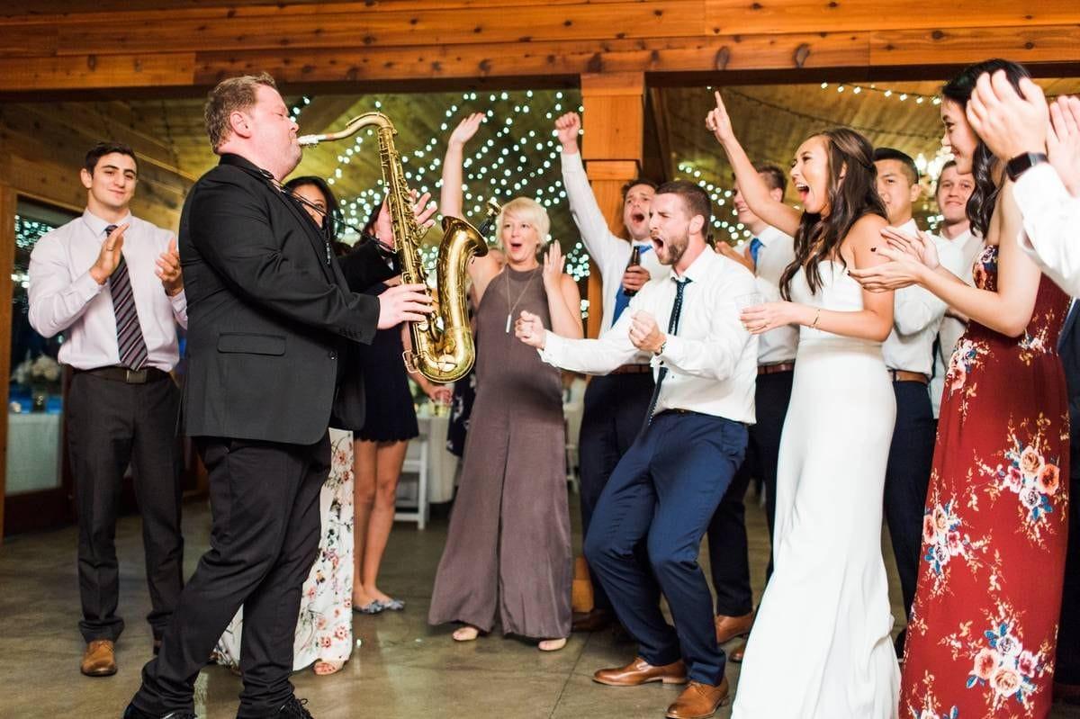A saxophonist in a black suit plays energetically to a cheering, clapping group of well-dressed people at an indoor event with string lights and wooden walls. Everyone looks joyful and engaged.