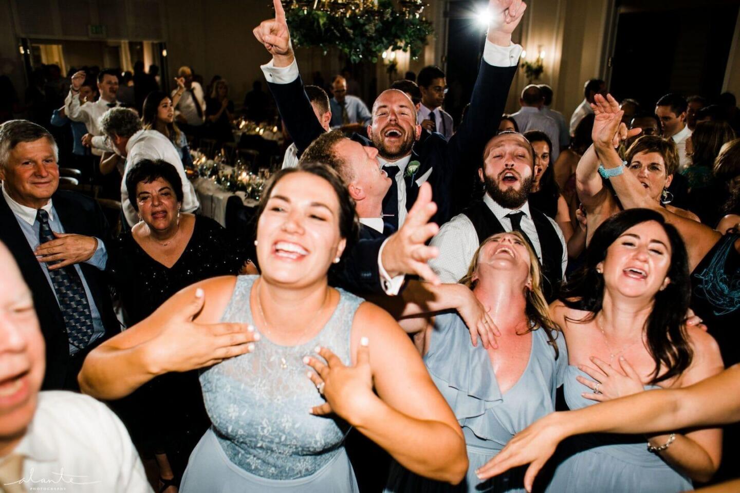 A joyful group of people, dressed in formal attire, laugh and cheer together on a crowded dance floor at a lively indoor event or wedding reception.