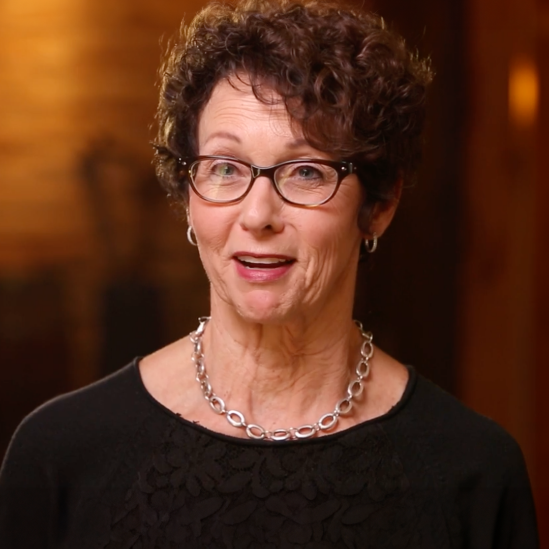 A middle-aged woman with short curly brown hair, glasses, and a silver necklace, wearing a black top, smiles while facing the camera. The background is softly blurred with warm lighting.