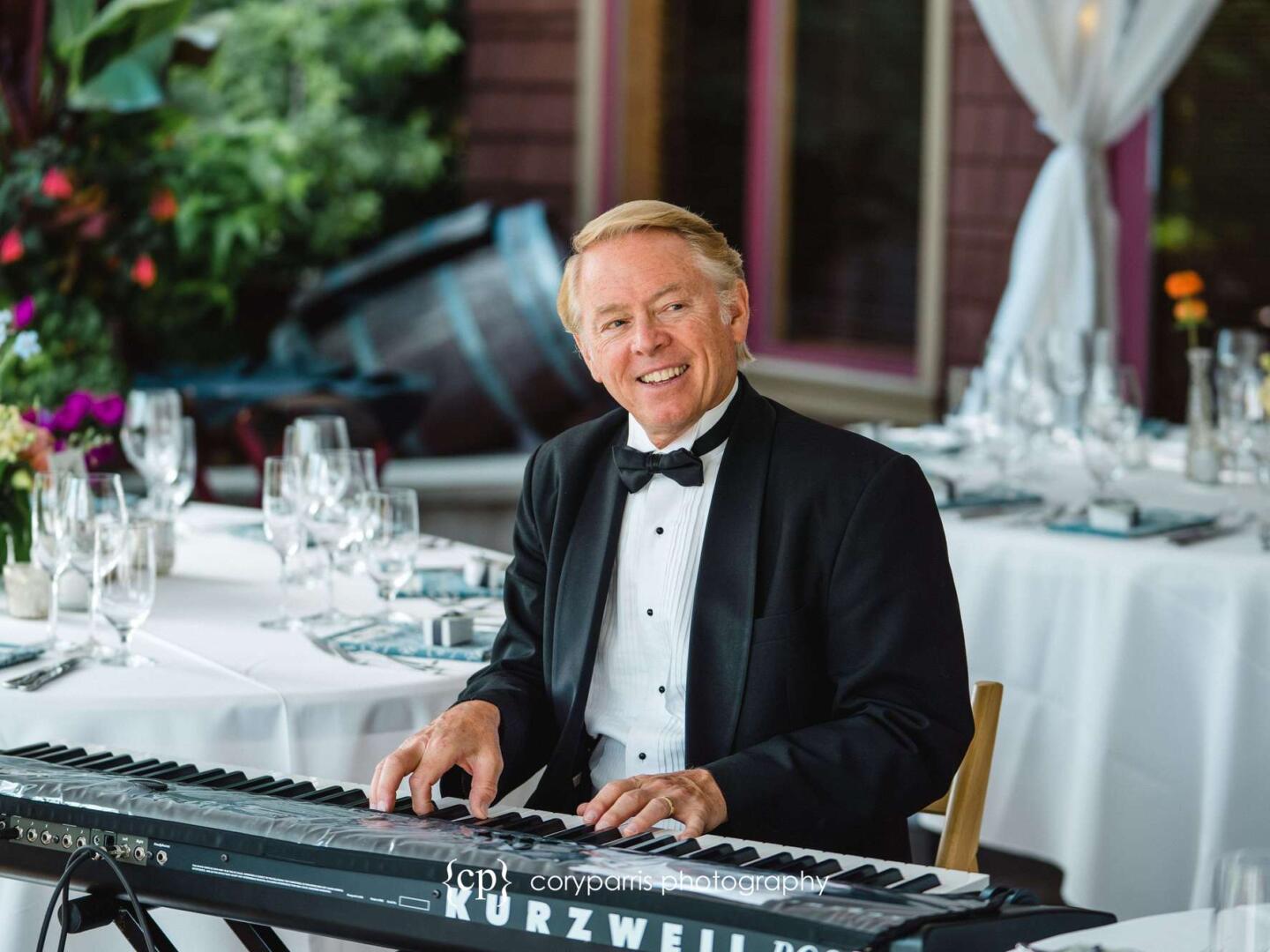 A man in a tuxedo smiles while playing a Kurzweil keyboard at a formal event with elegantly set tables and floral arrangements in the background.