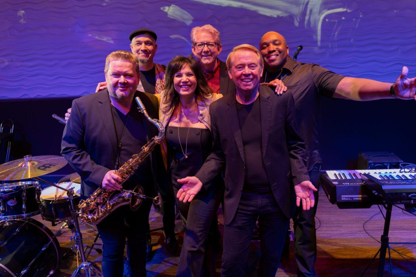 Six smiling musicians pose together on stage with instruments, including a saxophone, keyboard, and drum set, under purple stage lighting and a wavy backdrop.