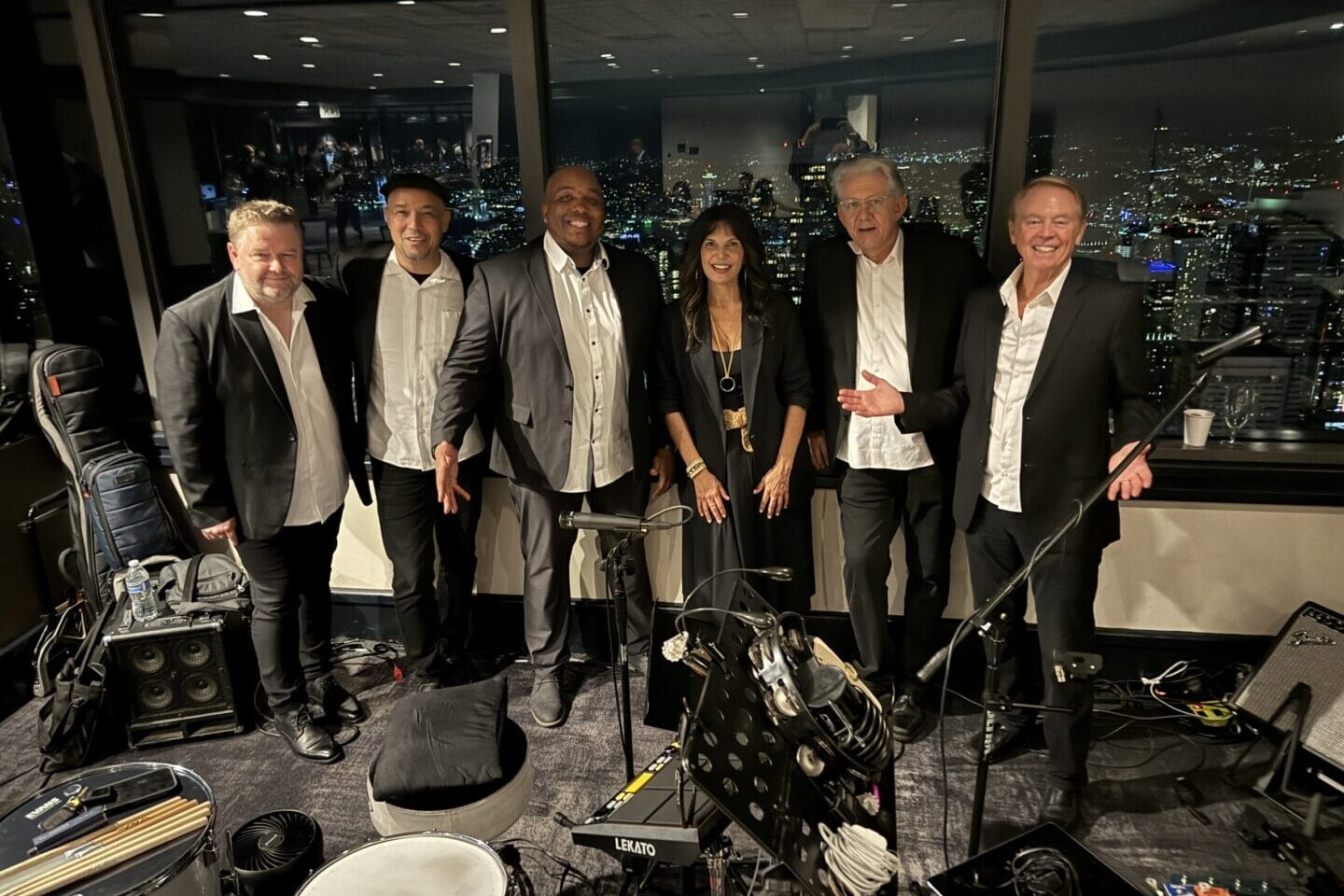 Six musicians, dressed in formal black and white clothing, stand together smiling in a city-view room at night, surrounded by musical instruments and equipment. Large windows reveal a brightly lit skyline behind them.
