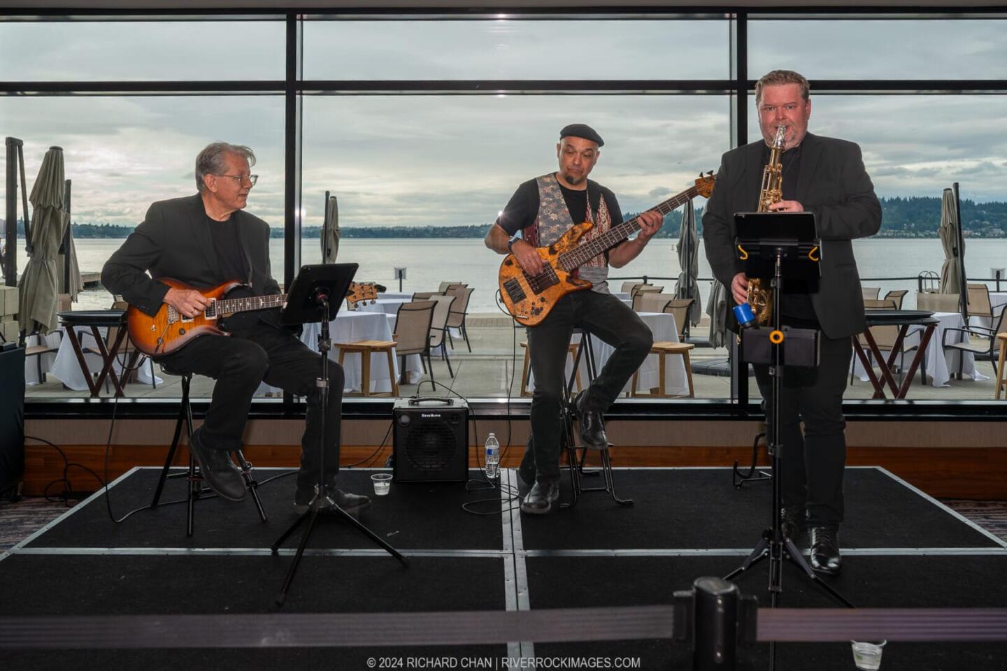 Three musicians perform indoors by large windows overlooking a waterfront. One plays electric guitar seated, another plays electric bass standing, and the third stands playing saxophone. Outdoor patio and water are visible in the background.
