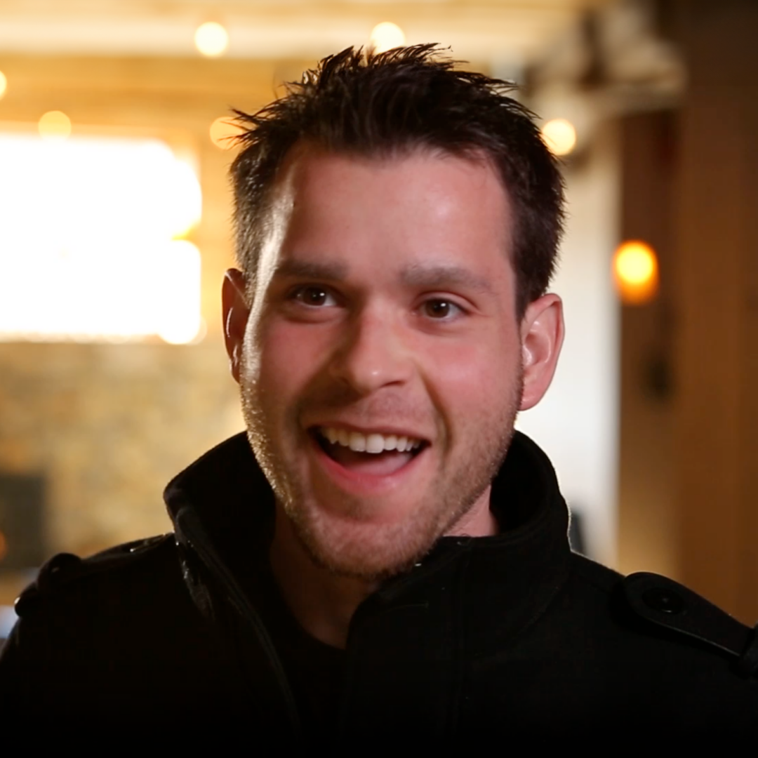 A smiling man with short dark hair and a trimmed beard wears a black jacket, standing indoors with warm lighting and blurred background lights.