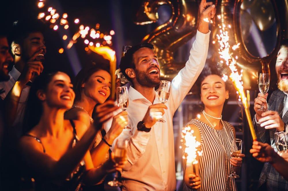 A group of people celebrate at a lively party, holding sparklers and champagne glasses. They are smiling and laughing, with gold balloons forming the number 2020 in the background.