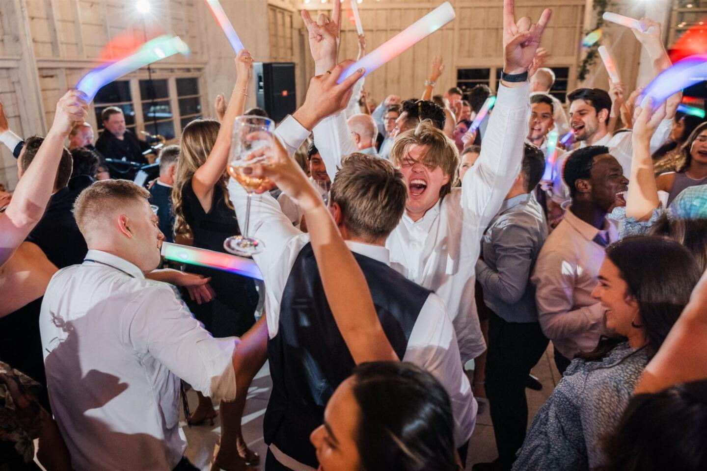 A group of people celebrate on a crowded dance floor, raising glow sticks and drinks in the air. Everyone appears excited and joyful, with one person in the center cheering enthusiastically.
