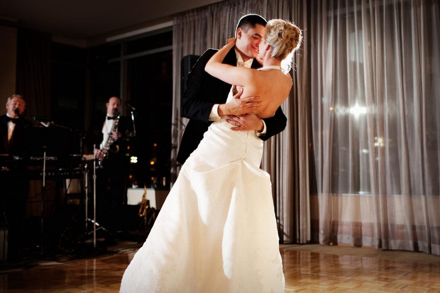 A bride and groom share a romantic first dance at their wedding reception, holding each other closely on a wooden dance floor while the wedding band performs a beautiful cover in the background.