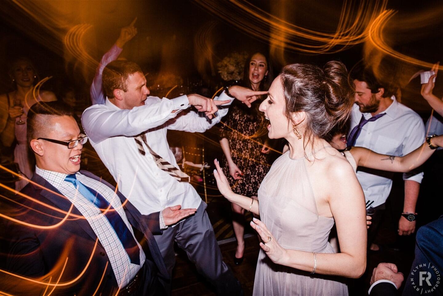 A group of people dance energetically at a party, laughing and interacting under swirling yellow lights, with one woman in a light dress and a man in a white shirt as focal points. The mood is joyful and celebratory.