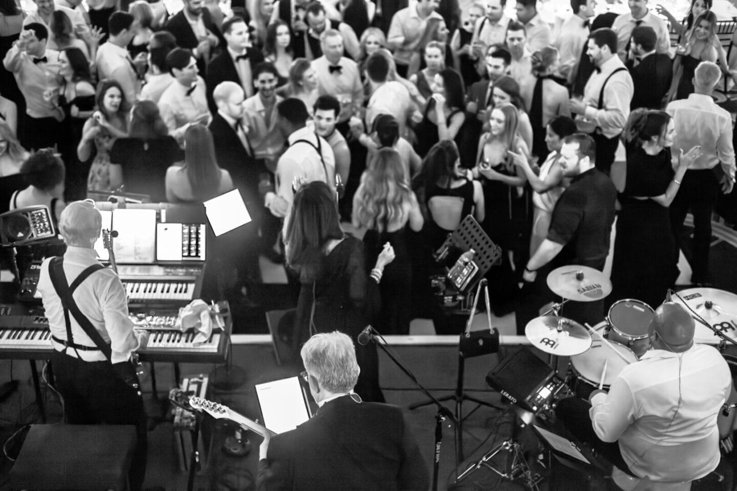 A black-and-white photo shows a live band performing on stage in front of a large, well-dressed crowd dancing and mingling at an indoor event or party.