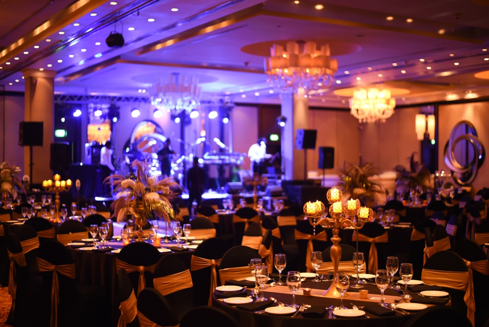 A lavish banquet hall set for an event, featuring round tables with black and gold decor, elegant chandeliers, candle centerpieces, and a lit stage with musical instruments in the background.