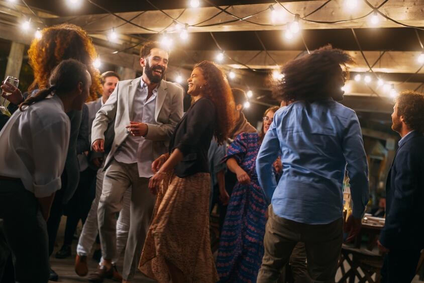 People dance and smile together at an outdoor party under string lights at night. The atmosphere is joyful and festive, with guests dressed in semi-formal attire and enjoying themselves.