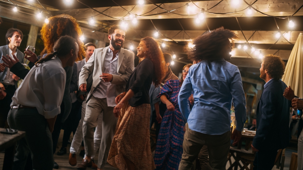 People dance and smile together at an outdoor party under string lights at night. The atmosphere is joyful and festive, with guests dressed in semi-formal attire and enjoying themselves.