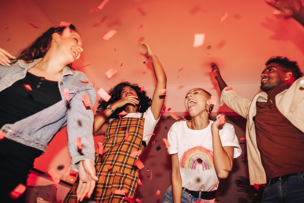 Four people joyfully dancing and laughing amid falling red confetti in a room with warm, pinkish lighting.